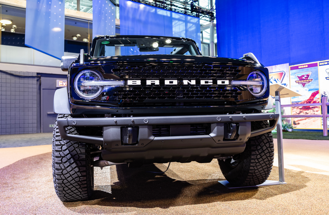 Front view of a black Ford Bronco with bold grille lettering and round LED headlights on display indoors, showcasing off-road tires and rugged bumper design