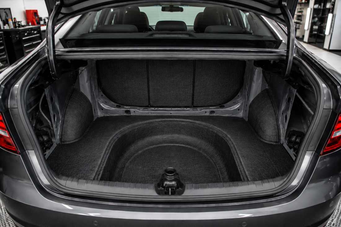 Open trunk of a modern sedan with trunk sound deadening installed across the trunk floor, spare tire well, and rear bulkhead using matte black material in a clean, brightly lit professional automotive workshop.