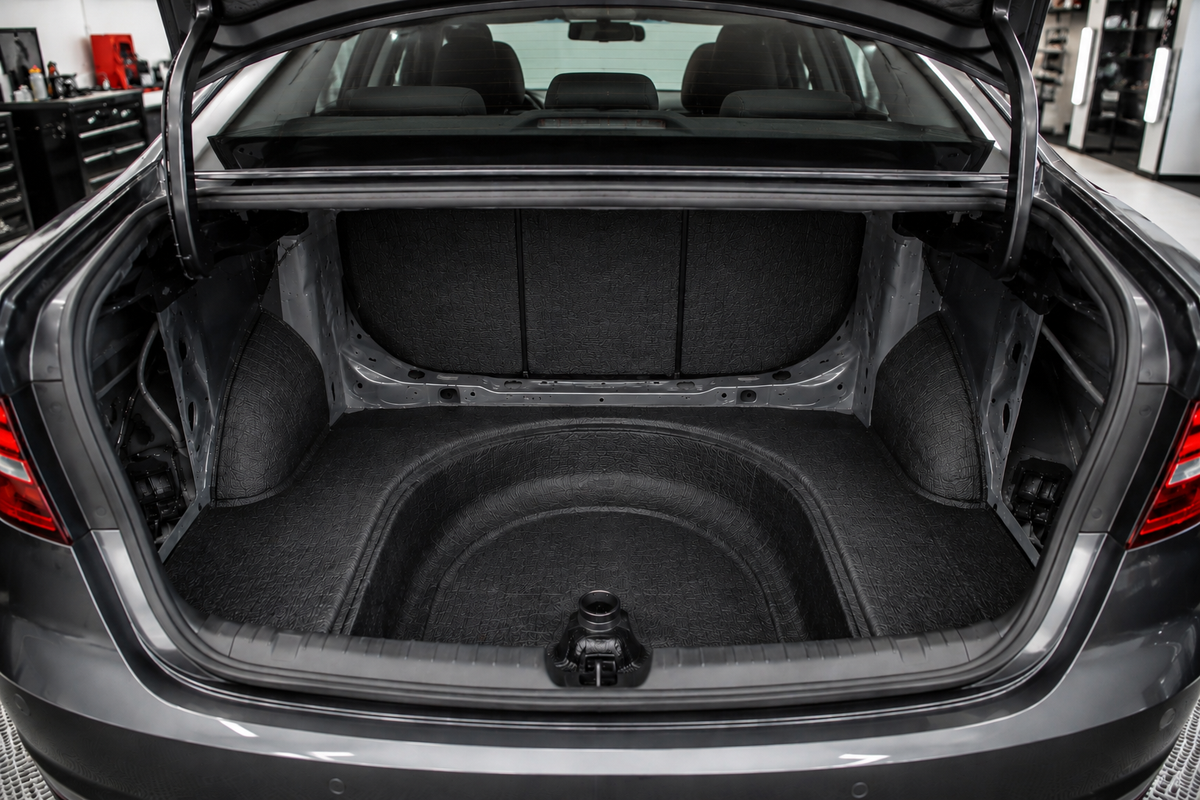 Open trunk of a modern sedan with trunk sound deadening installed across the trunk floor, spare tire well, and rear bulkhead using matte black material in a clean, brightly lit professional automotive workshop.
